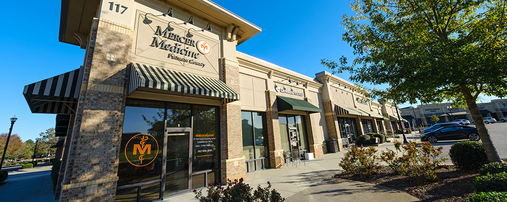 A tan brick building with a glass facade. A Mercer Medicine Putnam County sign hangs above the glass door with the Mercer Medicine logo to the left of the door.